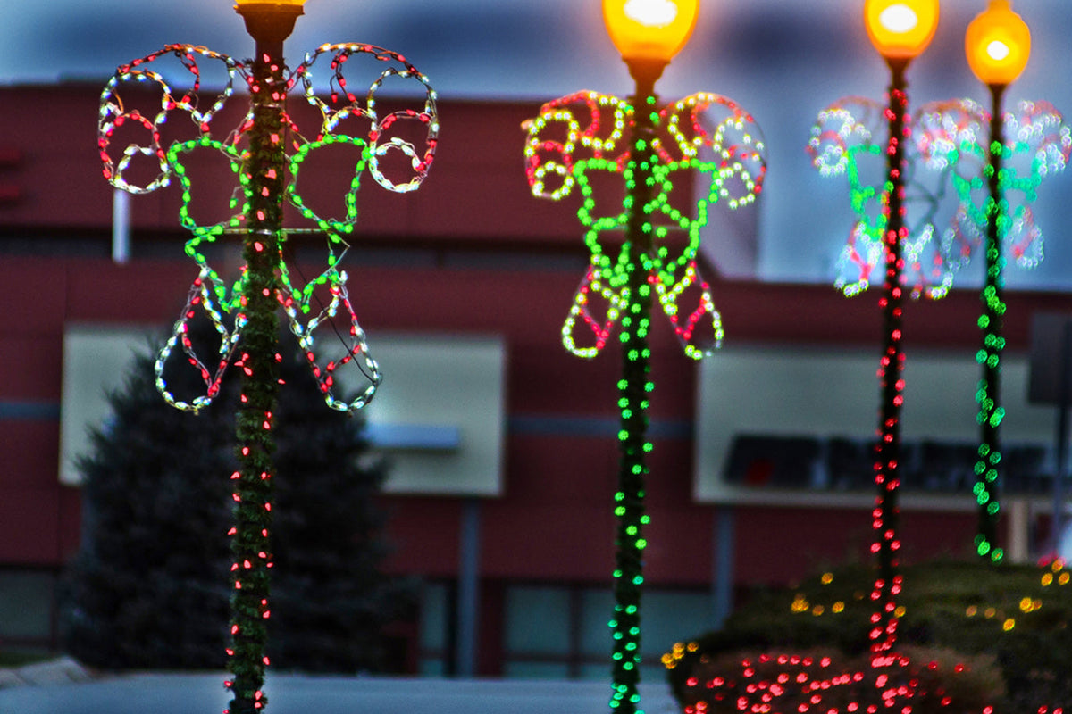 Outdoor holiday light decorations shaped like candy canes with green bows wrapped in red, green, and white lights, with glowing streetlamp posts in the background at dusk.