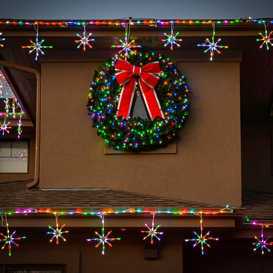 A large green Christmas wreath with a red ribbon bow is mounted on a house exterior, surrounded by multicolored string lights and hanging snowflake-shaped holiday lights.