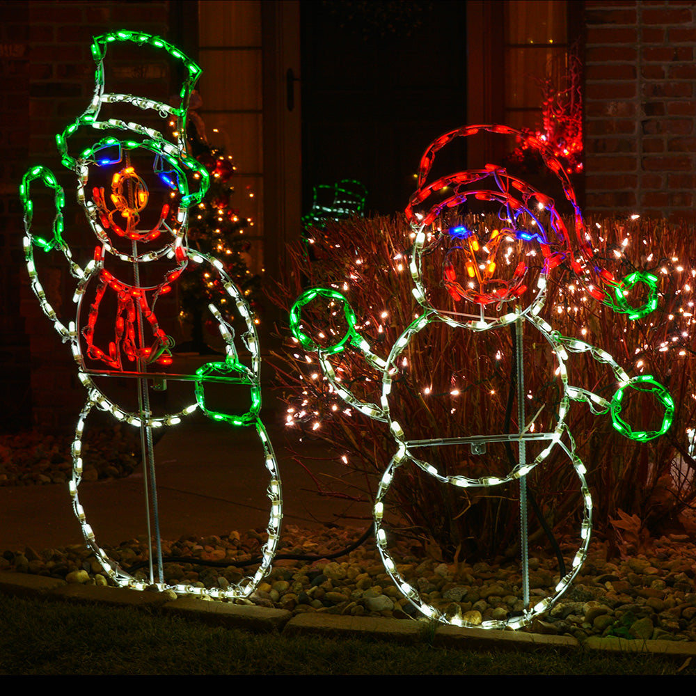 Two large outdoor snowman holiday light decorations illuminated at night in a front yard. One snowman is outlined in white with green and red accents, and the other is outlined in white with red and blue accents, set among shrubs with warm string lights.