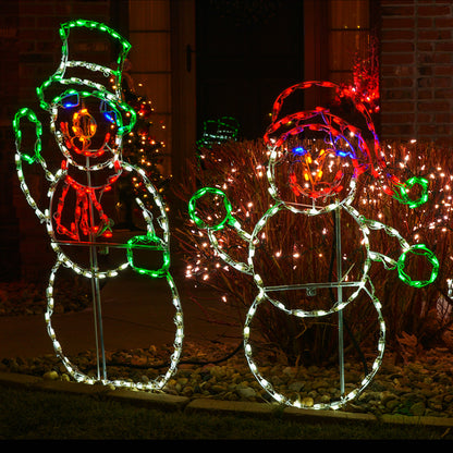 Two large outdoor snowman holiday light decorations illuminated at night in a front yard. One snowman is outlined in white with green and red accents, and the other is outlined in white with red and blue accents, set among shrubs with warm string lights.